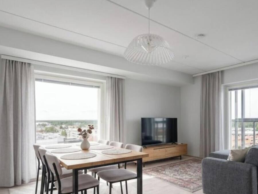 A modern living room with a dining area, large windows, a flat-screen TV, and a grey sofa.
