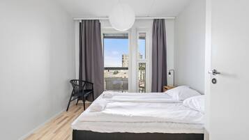 Un dormitorio con cama, una silla, un escritorio y vistas a la ciudad a través de amplios ventanales.