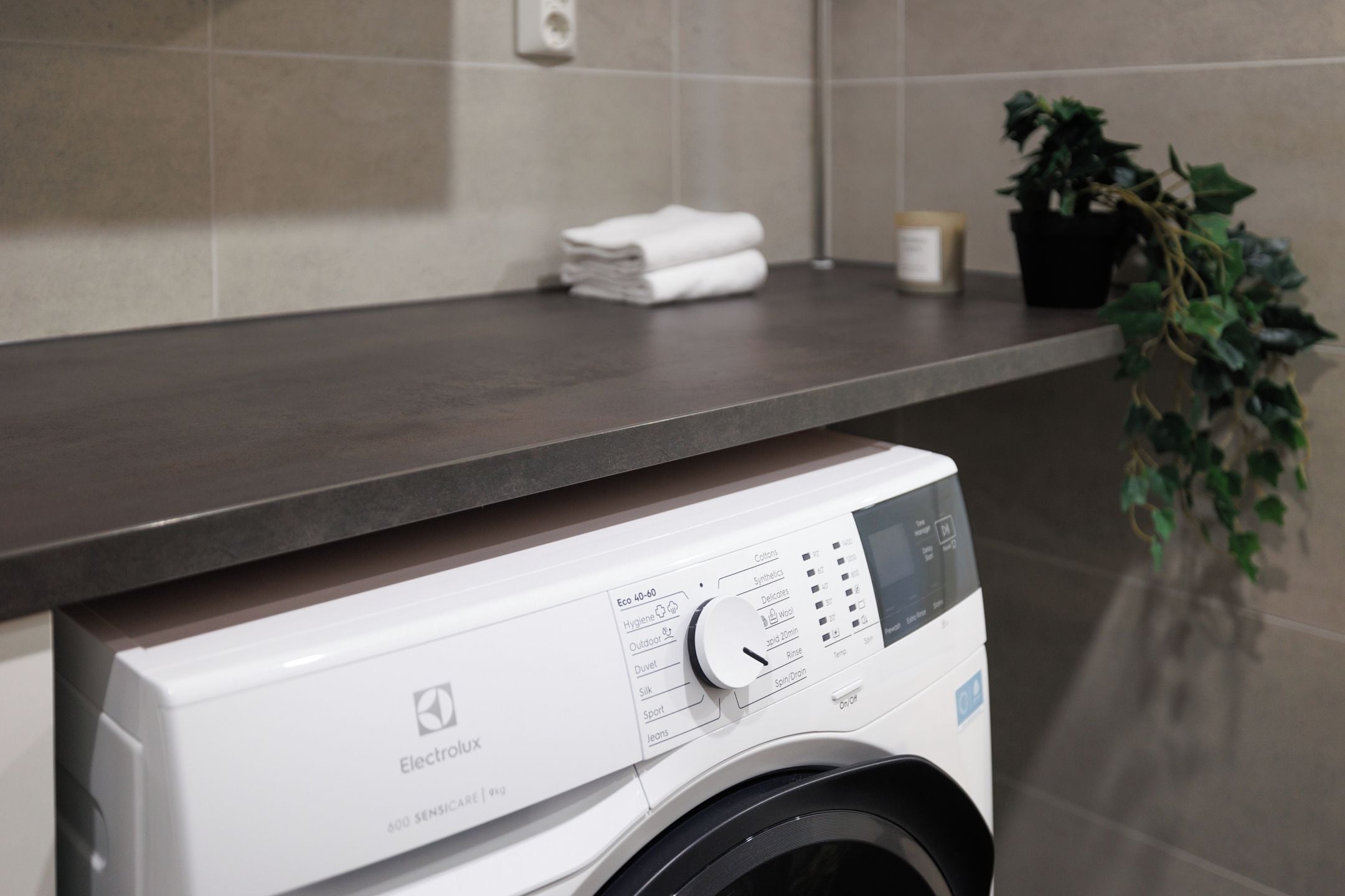 A front-loading washing machine with a control panel, placed in a modern bathroom with a grey countertop and a potted plant.