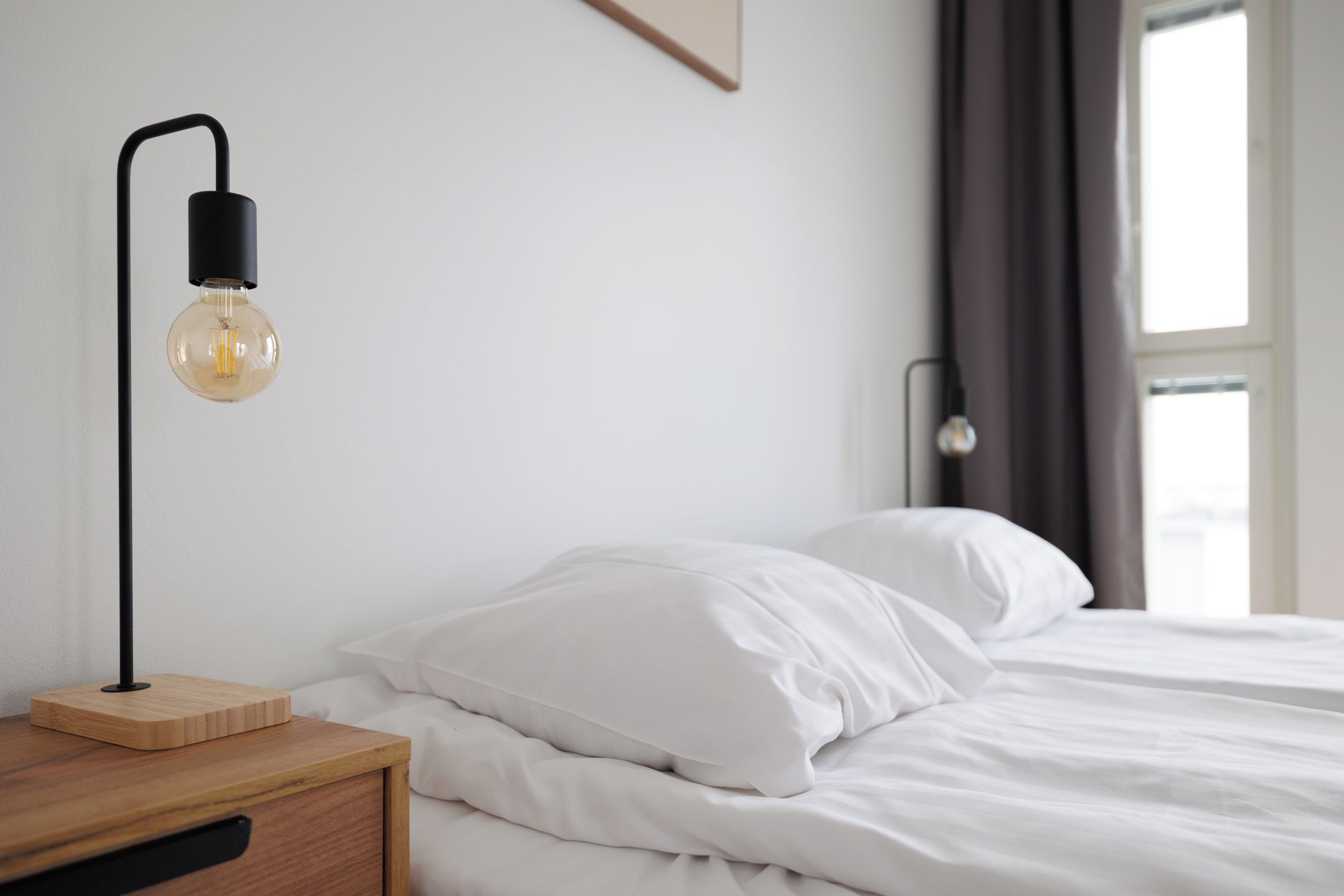 Cama bem arrumada com lençóis brancos, mesa de cabeceira de madeira com abajur preto e janela com cortinas.