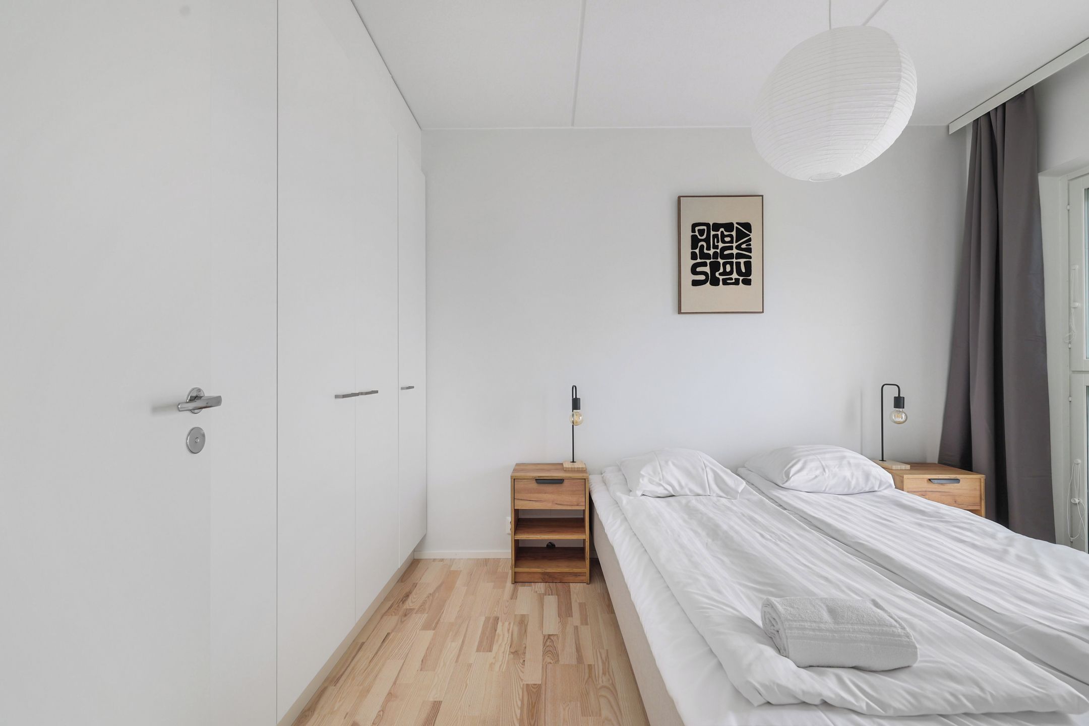 A minimalist bedroom with a bed, bedside tables, a hanging light fixture, and a wall-mounted artwork.
