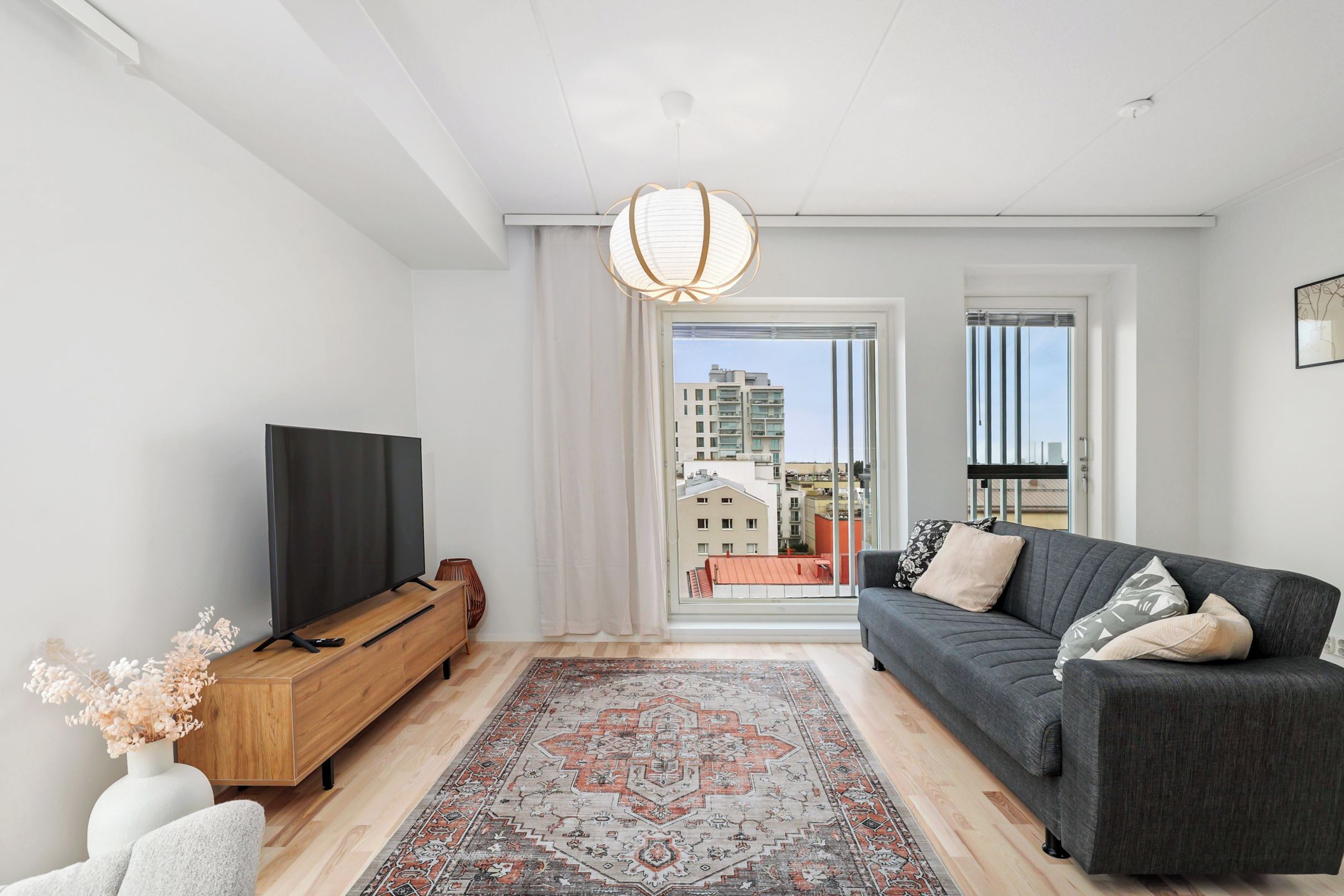 Un salon moderne doté d’un téléviseur à écran plat, d’un canapé foncé, d’un tapis à motifs et offrant une vue sur la ville grâce à de grandes fenêtres.