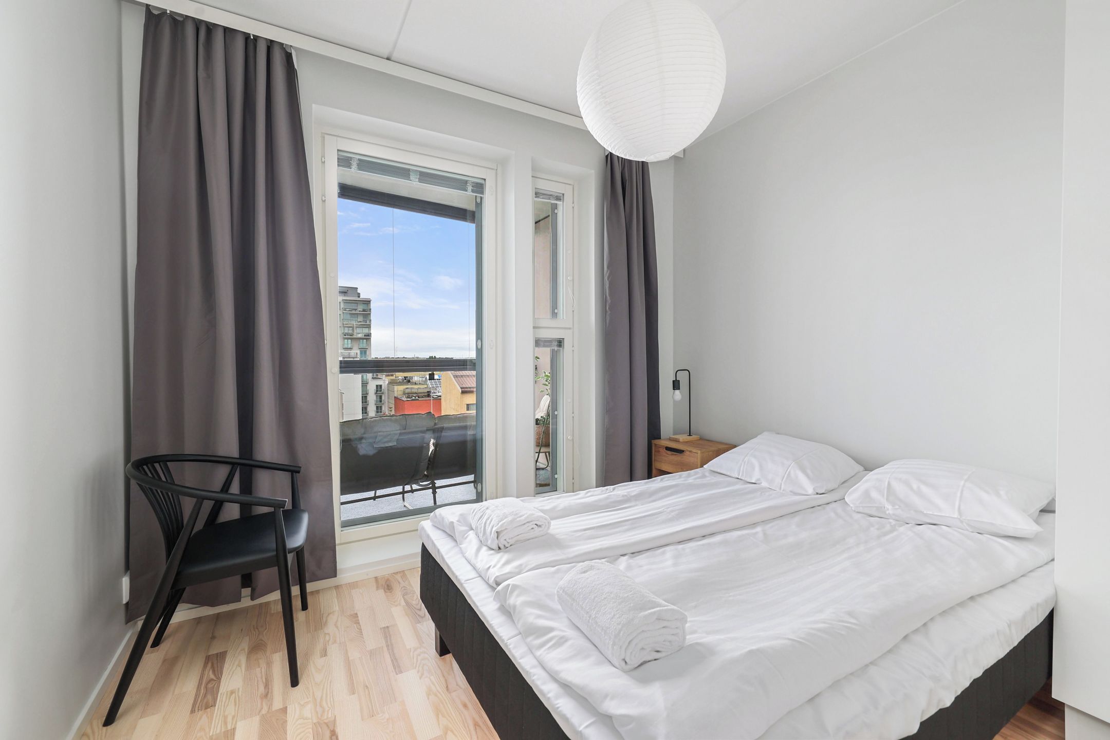 A bedroom with a bed, a chair, a nightstand, a lamp, and a view of the city through a window.