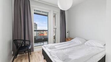 A bedroom with a bed, a chair, a nightstand, a lamp, and a view of the city through a window.