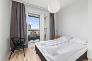 Un dormitorio con cama, una silla, una mesita de noche, una lámpara y vistas a la ciudad a través de una ventana.
