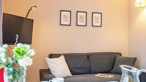 Ein Zimmer mit grauem Sofa, einem Flachbildfernseher, gerahmten Kunstwerken an der Wand und einer Vase mit Blumen.