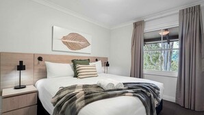 A hotel room with a bed, a nightstand, a wall-mounted lamp, a window with curtains, and a view of trees outside. - Villa Boronia - 2 Bedroom Villa at Cypress Lakes Resort (POKOLBIN)