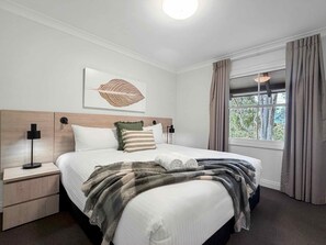 Ein Hotelzimmer mit einem Bett, einem Nachttisch, einer Wandlampe, einem Fenster mit Vorhängen und Blick auf Bäume.