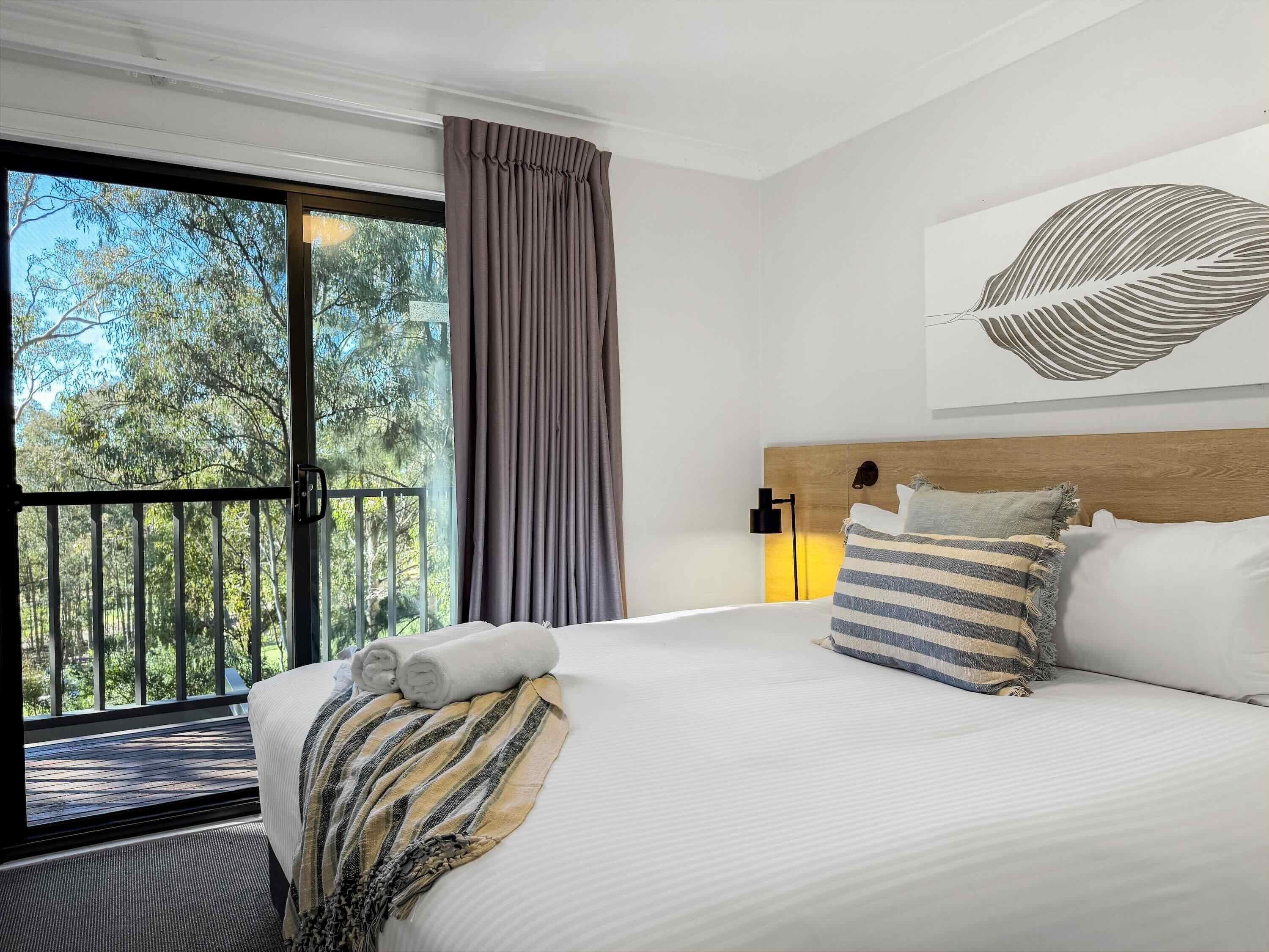 A hotel room with a large bed, a view of trees through a balcony, and a modern headboard.