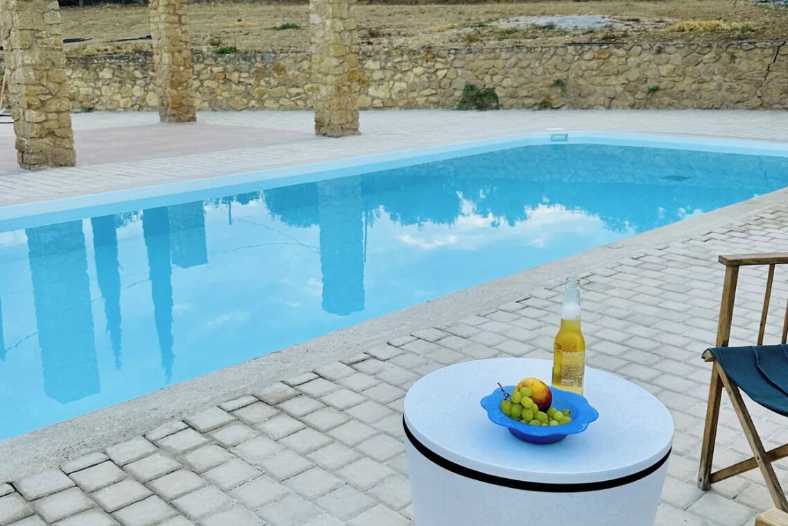 A swimming pool with a stone wall backdrop, a round table with a fruit bowl and a bottle, and a chair.