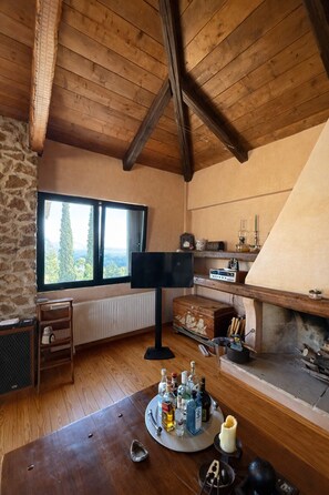A cozy room with a stone wall, wooden ceiling beams, a large window, a fireplace, and a table with bottles. - Olive House Villa Galaxidi (Galaxidi)