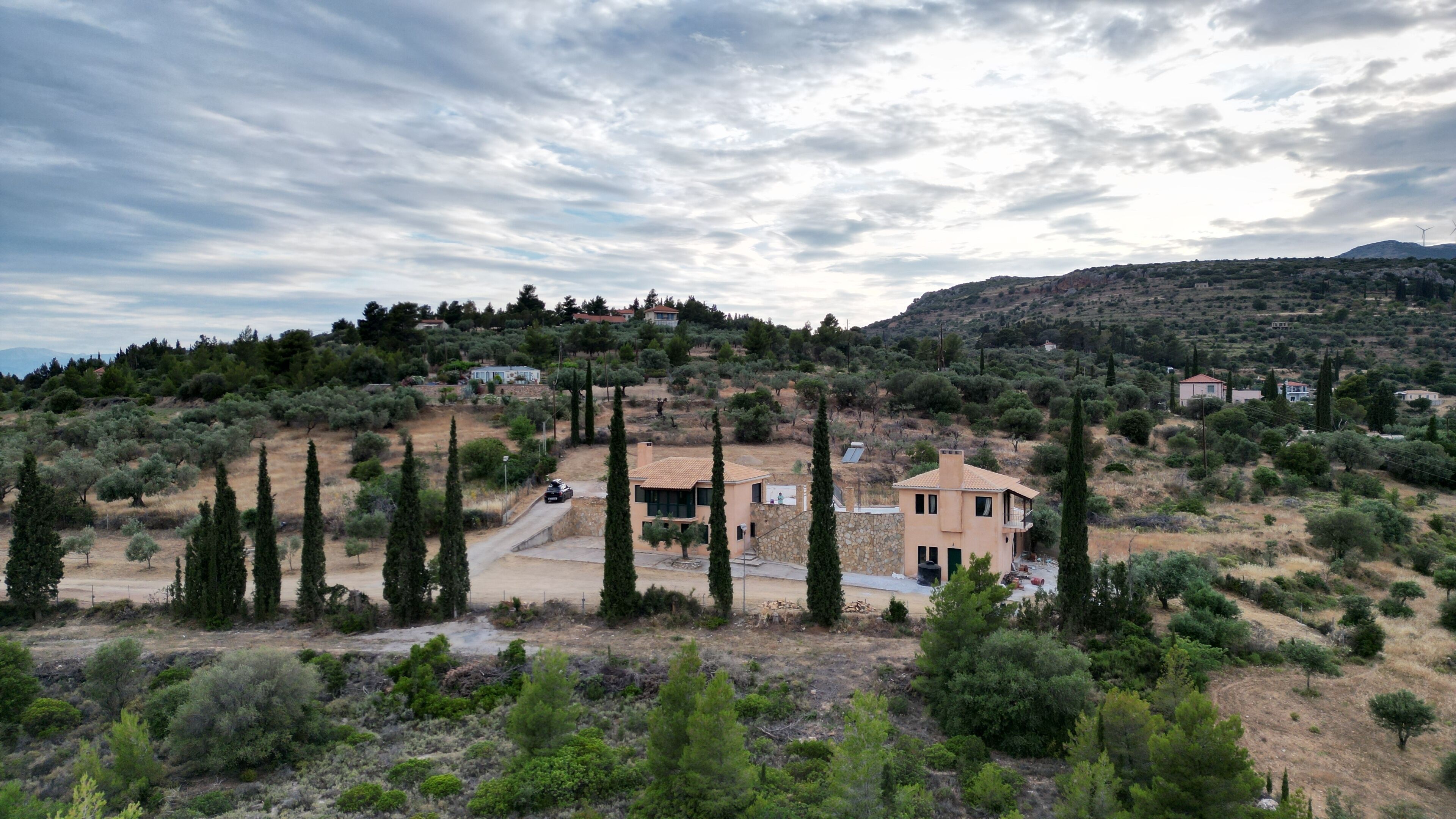 A rural landscape with a house surrounded by trees and a hilly backdrop.