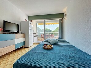 Ein Schlafzimmer mit Bett, Fernseher, Kühlschrank und einem Balkon mit Gartenmöbeln.