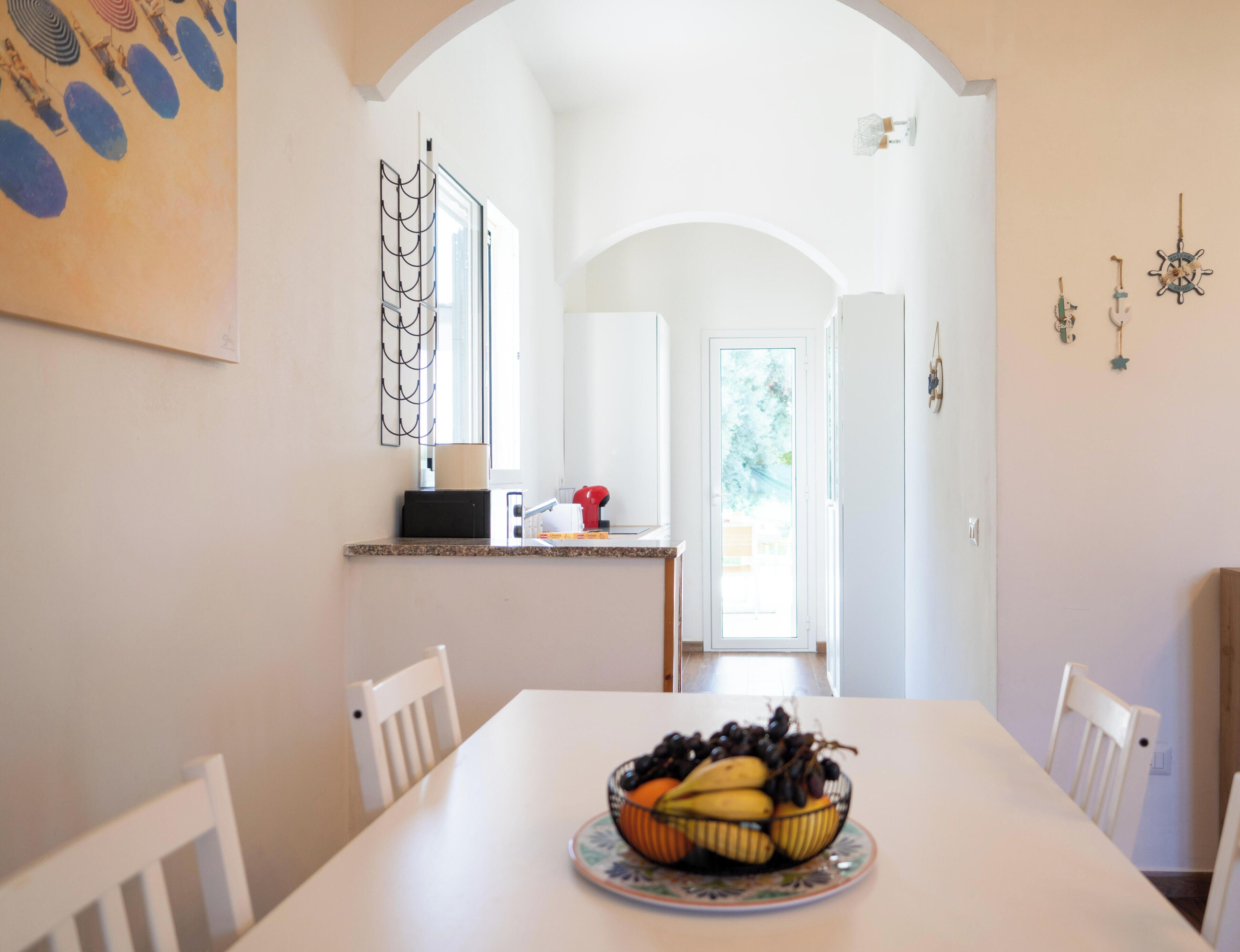Un coin repas avec une table blanche, des chaises et un saladier de fruits. Il y a également un coin cuisine avec une machine à café et une fenêtre.