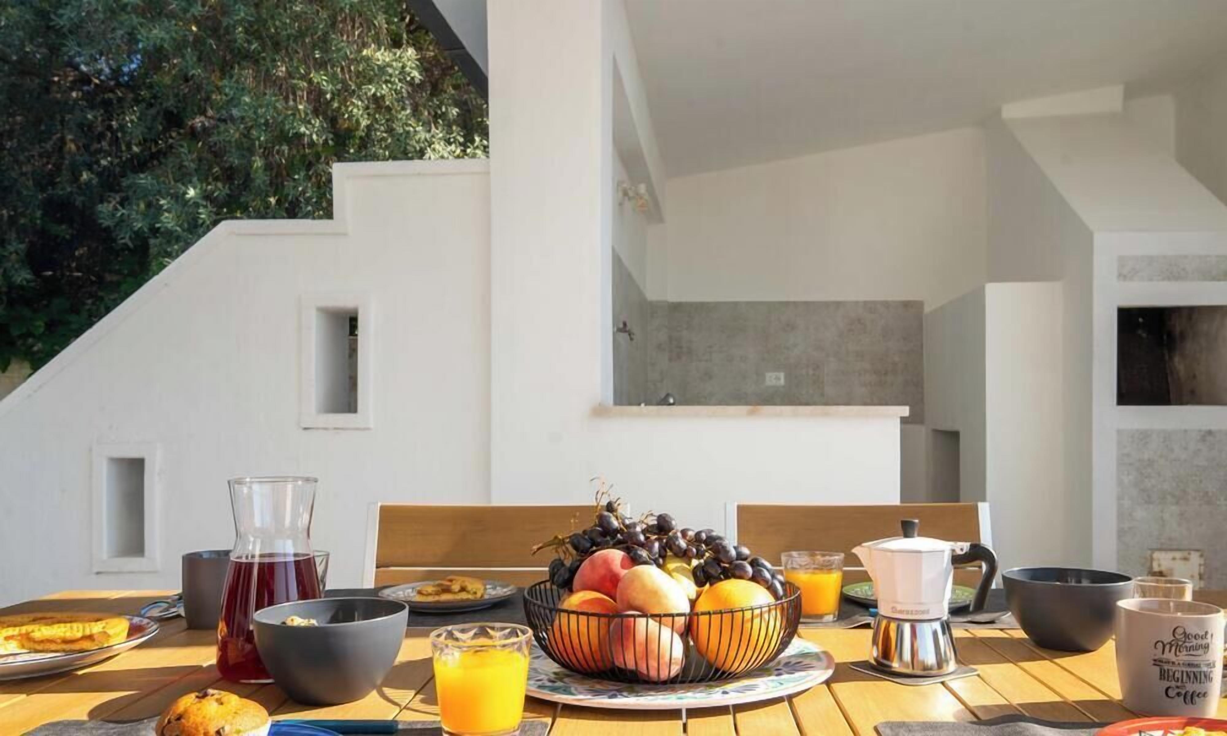 A breakfast table set with fruits, coffee, and juice in a modern outdoor setting with a staircase and greenery in the background.