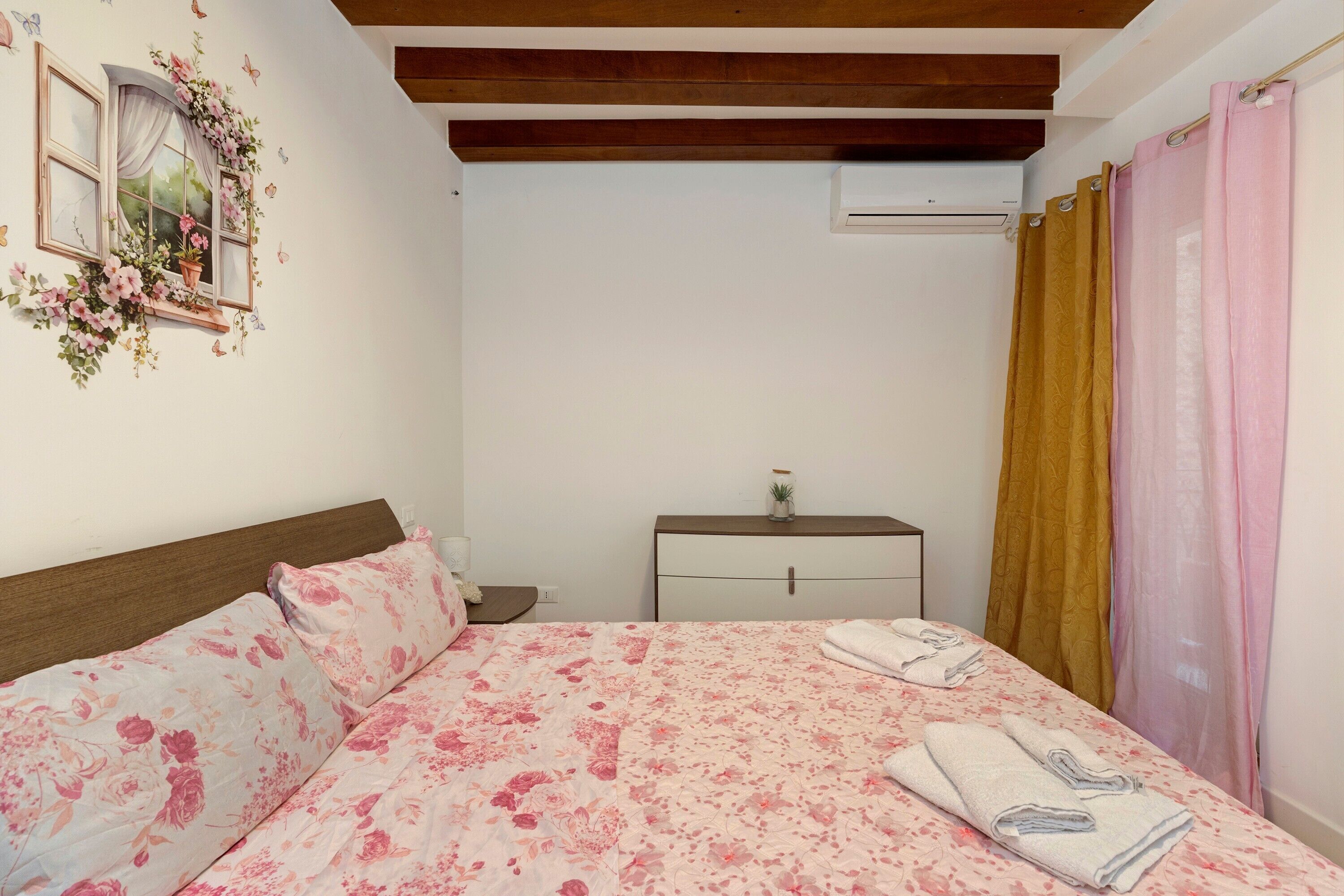 Ein Schlafzimmer mit einem Bett, einem Nachttisch, einer kleinen Kommode, einer Klimaanlage und einem Fenster mit Vorhängen.