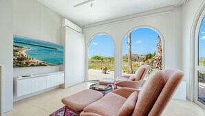 Un soggiorno moderno con una grande televisione a schermo piatto, posti a sedere comodi e vista su una spiaggia e palme.