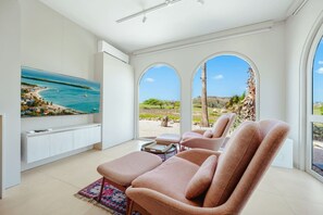 Ein modernes Wohnzimmer mit großem Flachbildfernseher, bequemen Sitzgelegenheiten und Blick auf Strand und Palmen.