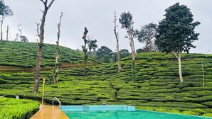 A swimming pool with a wooden deck, surrounded by terraced tea plantations and trees. - Hotel Wayanad gate (Vythiri)