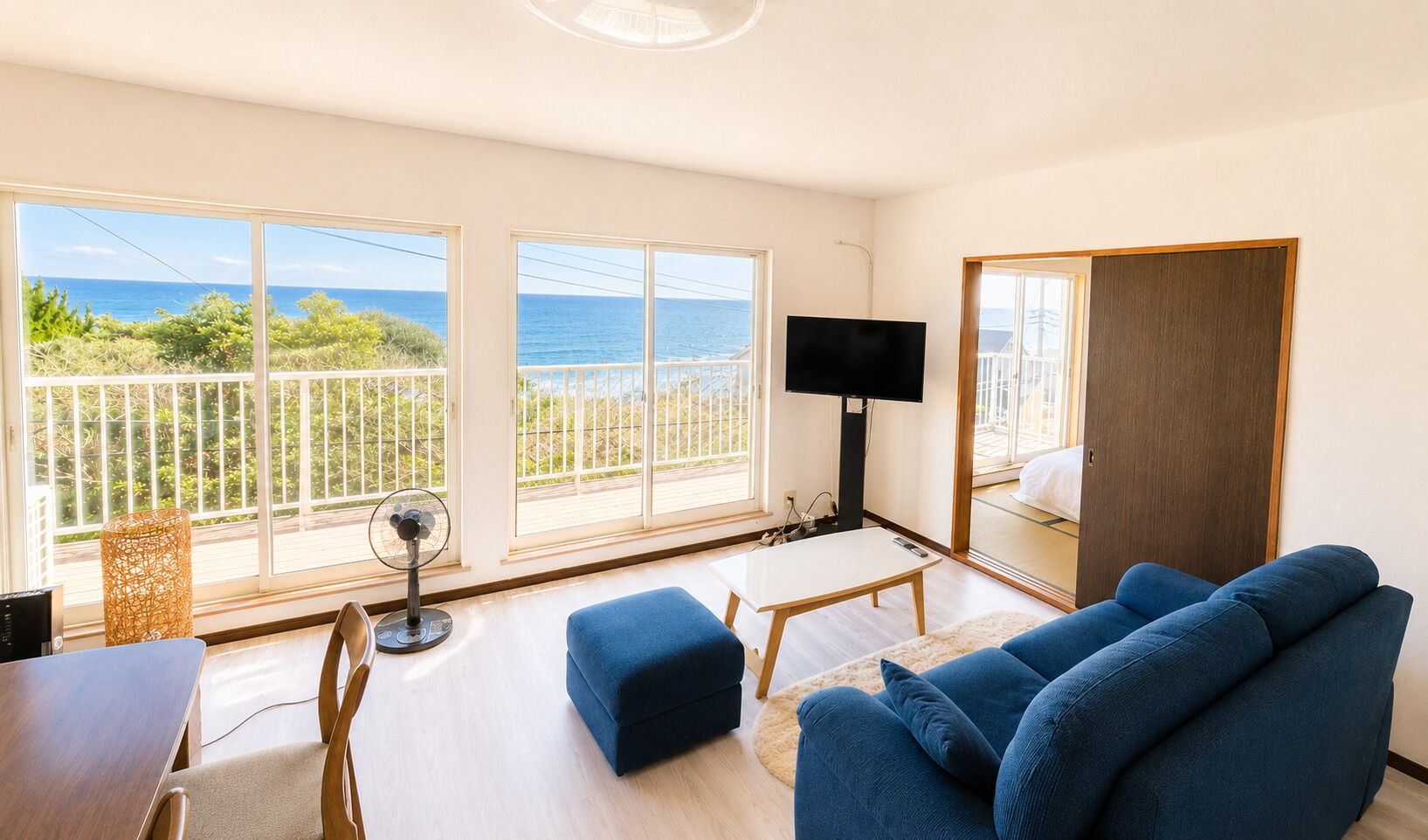 Un salon moderne avec un grand téléviseur, des canapés bleus, une table à manger et un balcon offrant une vue sur la mer.