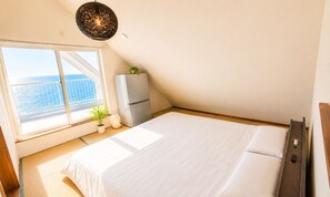 A bedroom with a large bed, a window with a view of the sea, a refrigerator, and a bedside table.