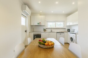 A modern kitchen with white cabinetry, a dining table with a fruit bowl, and a window with a view. - Cascais Boutique Retreat by Clama Retreats (Cascais)