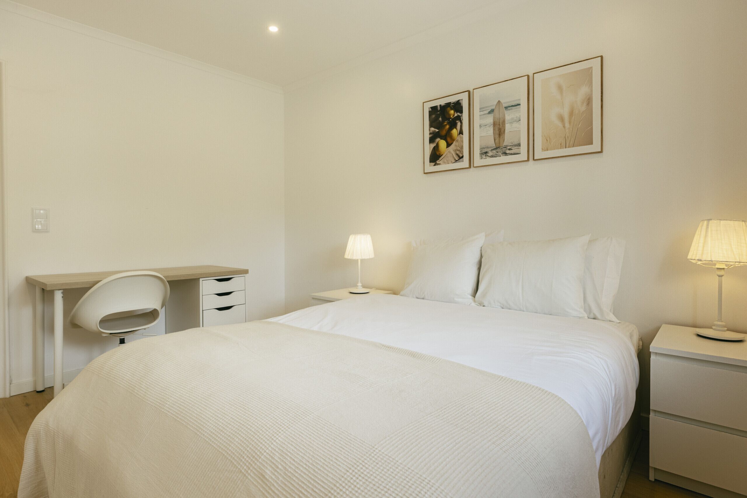 Un dormitorio minimalista con cama, mesitas de noche, un escritorio y dos cuadros colgados en la pared.