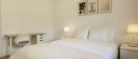 Un dormitorio minimalista con cama, mesitas de noche, un escritorio y dos cuadros colgados en la pared.