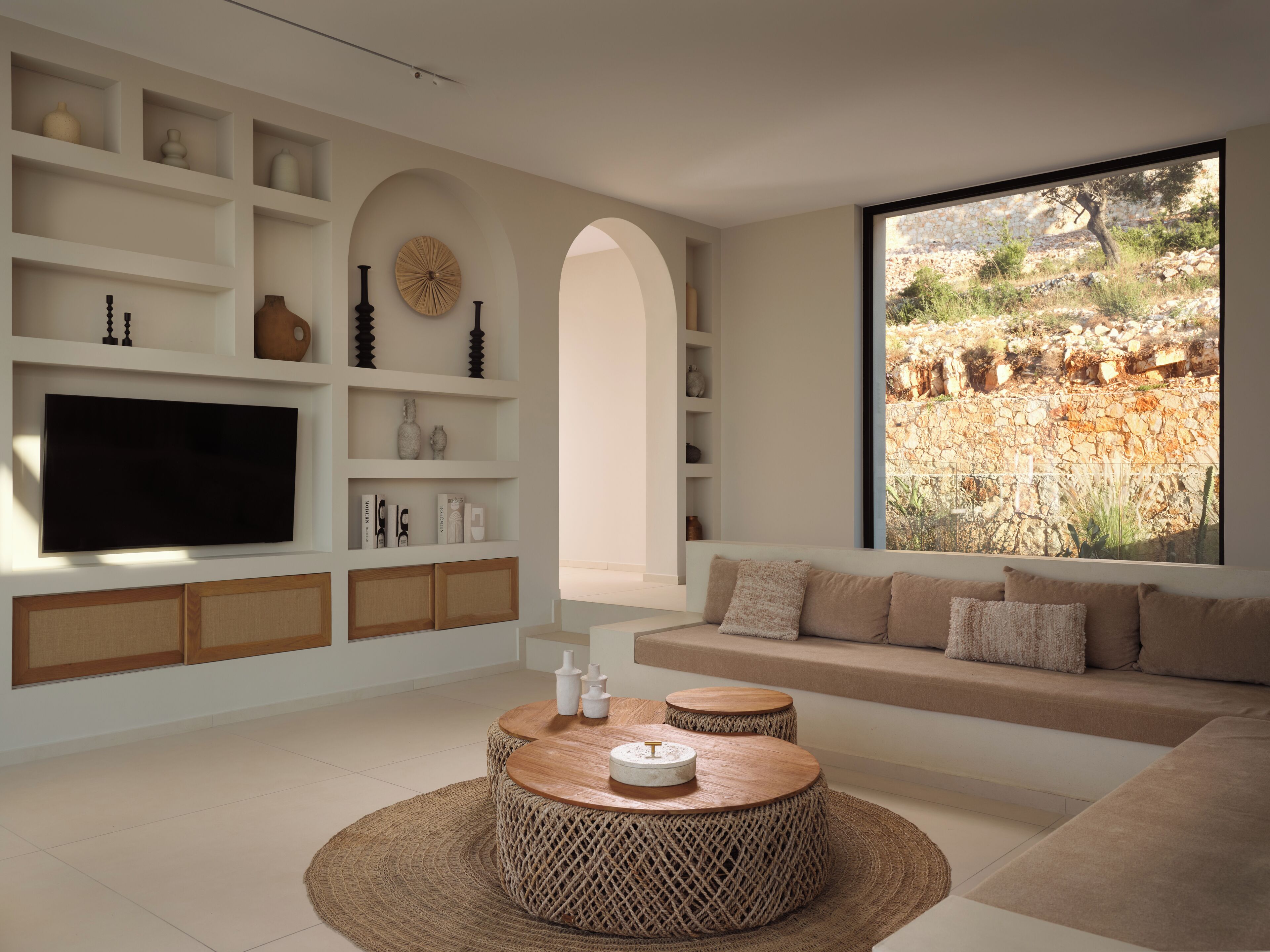 Ein modernes Wohnzimmer mit einem großen Fernseher, eingebauten Regalen, einem runden Couchtisch und Blick auf eine felsige Landschaft.