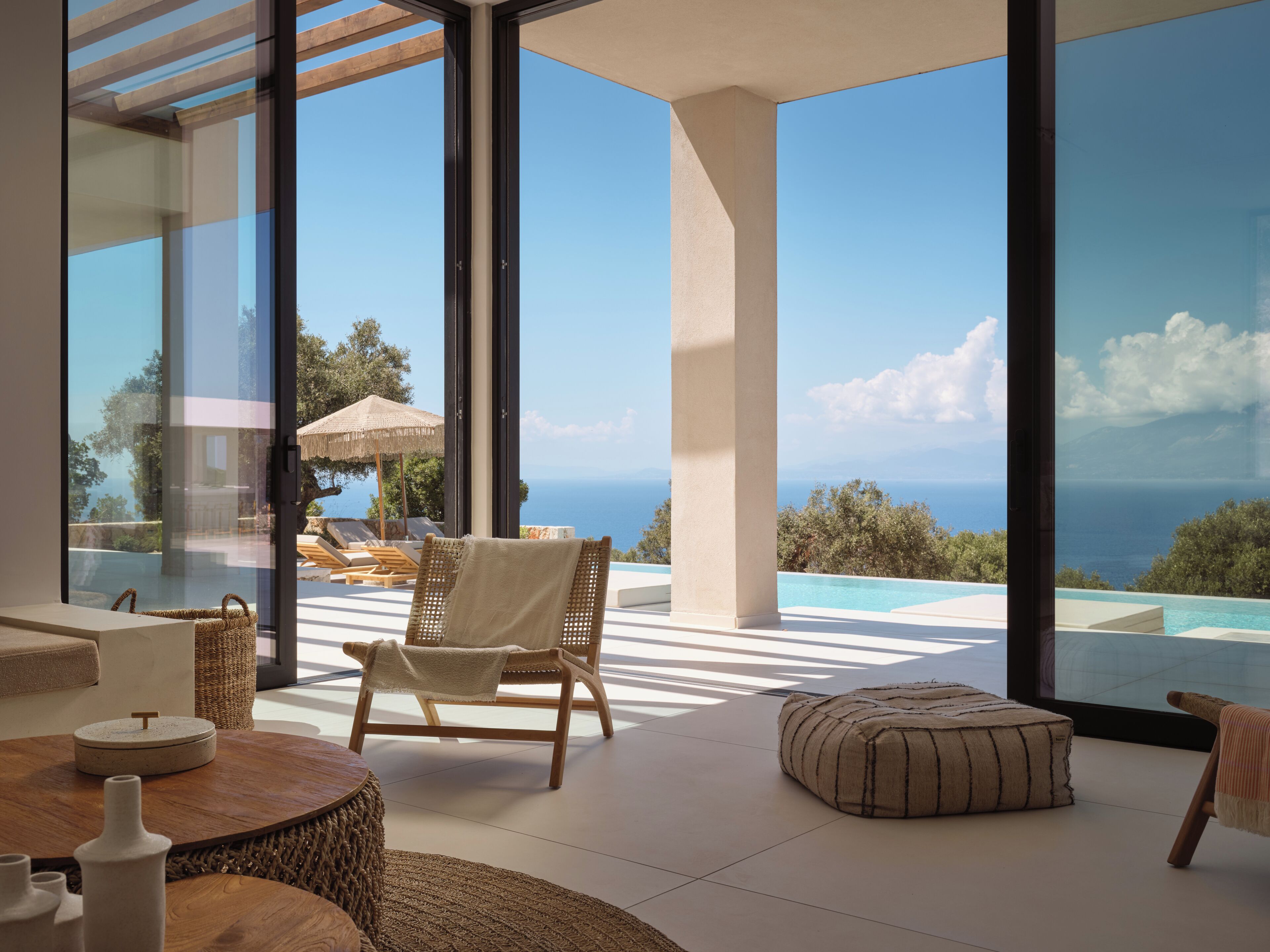 Un interior modern amb vistes al mar, que inclou una zona d'estar amb una cadira de vimet i un otomà, una taula rodona de fusta i una finestra gran.