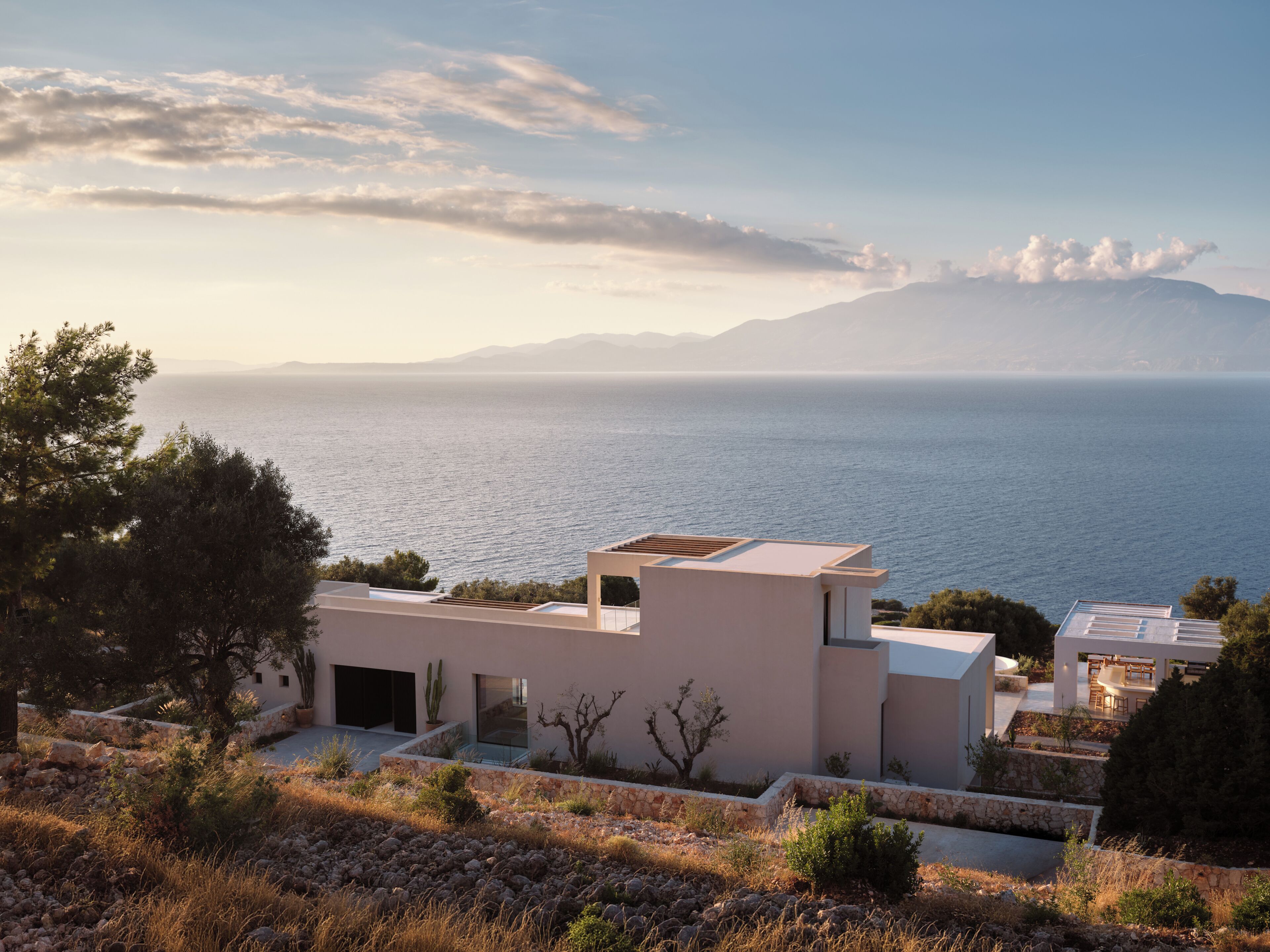 A modern building with a flat roof, situated on a hillside overlooking a body of water and distant mountains.