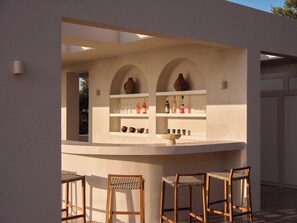 A bar area with a counter, shelves with bottles, glasses, and a bowl. There are three wooden chairs with woven seats. - Luxury 6-bedroom villa in north Zakynthos | Private pool & sea views (Korithi)