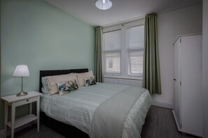 Een slaapkamer met een bed, een nachtkastje met een lamp, een raam met gordijnen en een deur.