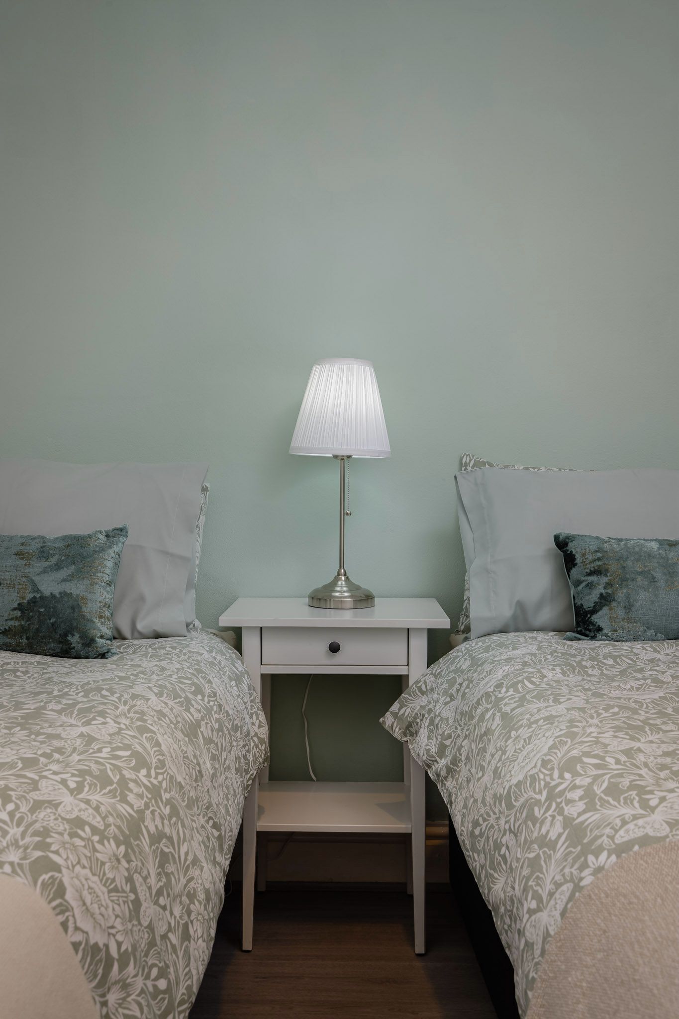 A bedroom with two beds, a bedside table with a lamp, and a drawer.
