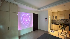 Ein modernes Wohnzimmer mit einem an der Wand montierten Projektor, der eine Herzanimation zeigt, einem Küchenbereich mit Schränken und einer Arbeitsplatte sowie einer Tür zu einem weiteren Raum.