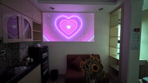 Una habitación con un proyector de pared que muestra una animación en forma de corazón, un sofá, un jarrón con girasoles y un mueble con espejo.