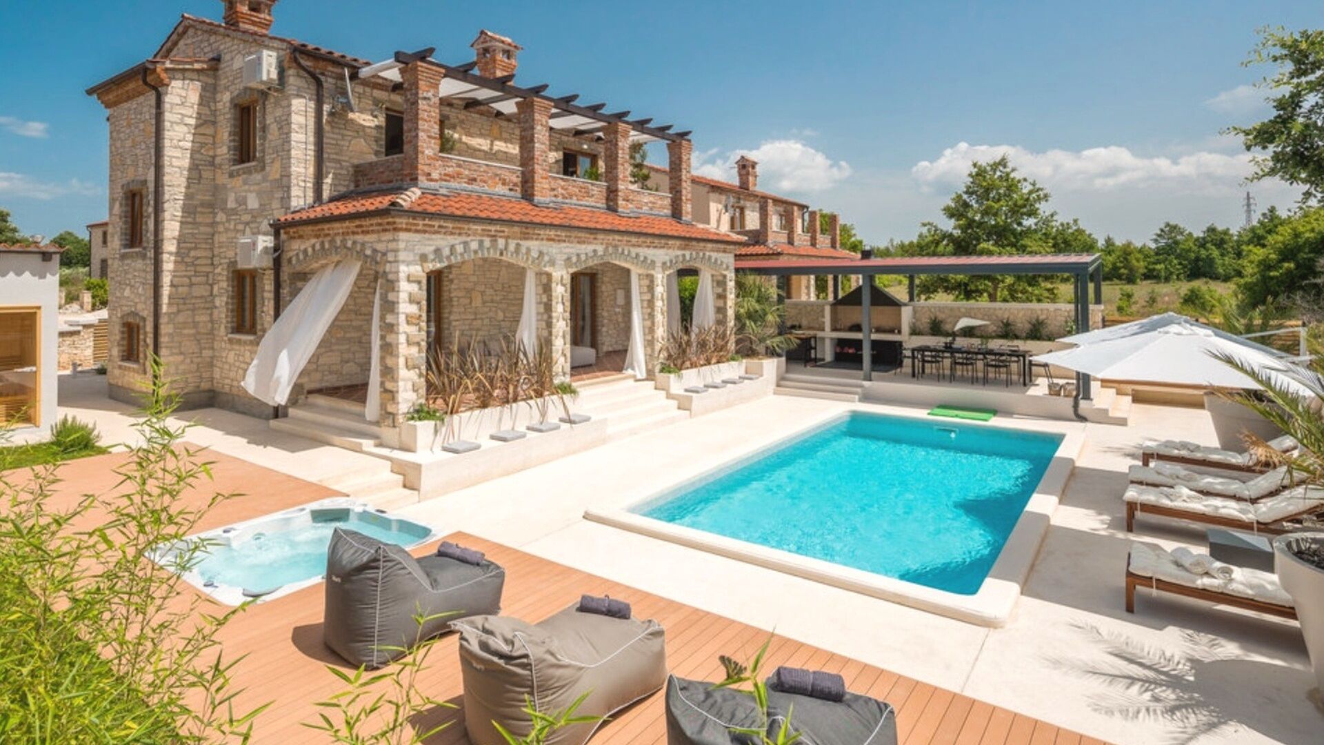 A pool area with a hot tub, lounge chairs, and a stone building with a tiled roof.