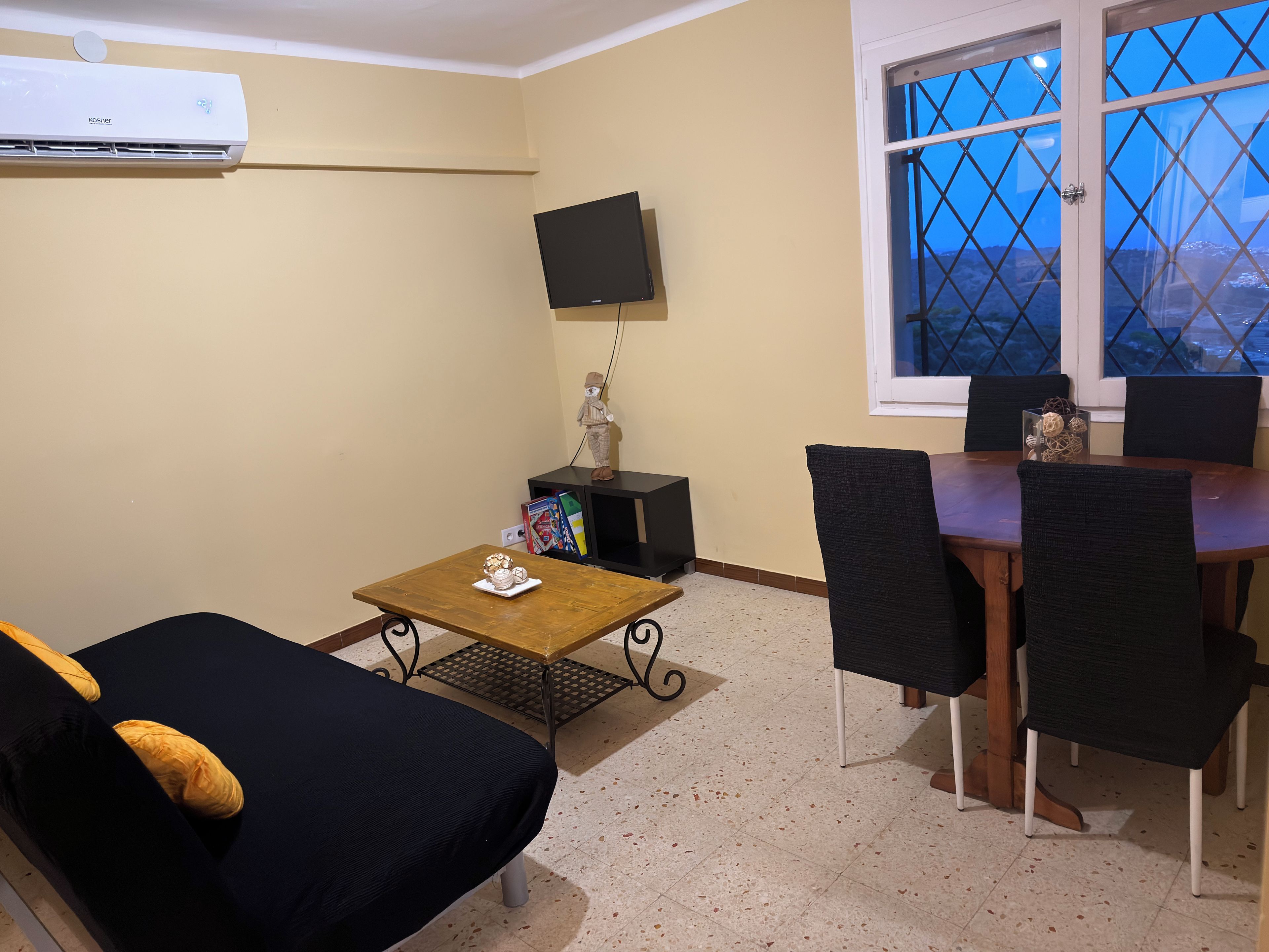 A room with a bed, a small table with a decorative item, a TV mounted on the wall, and a window with a view.