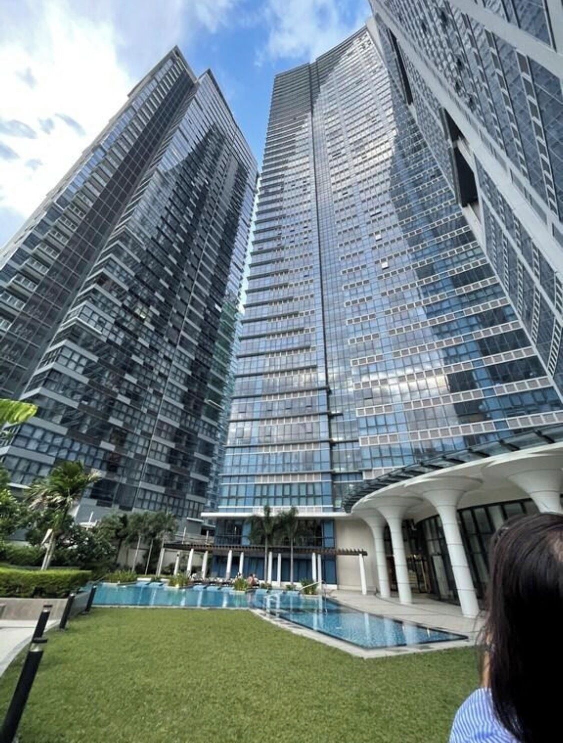 A modern high-rise building with a glass facade, a pool area with a white structure, and a well-maintained lawn.