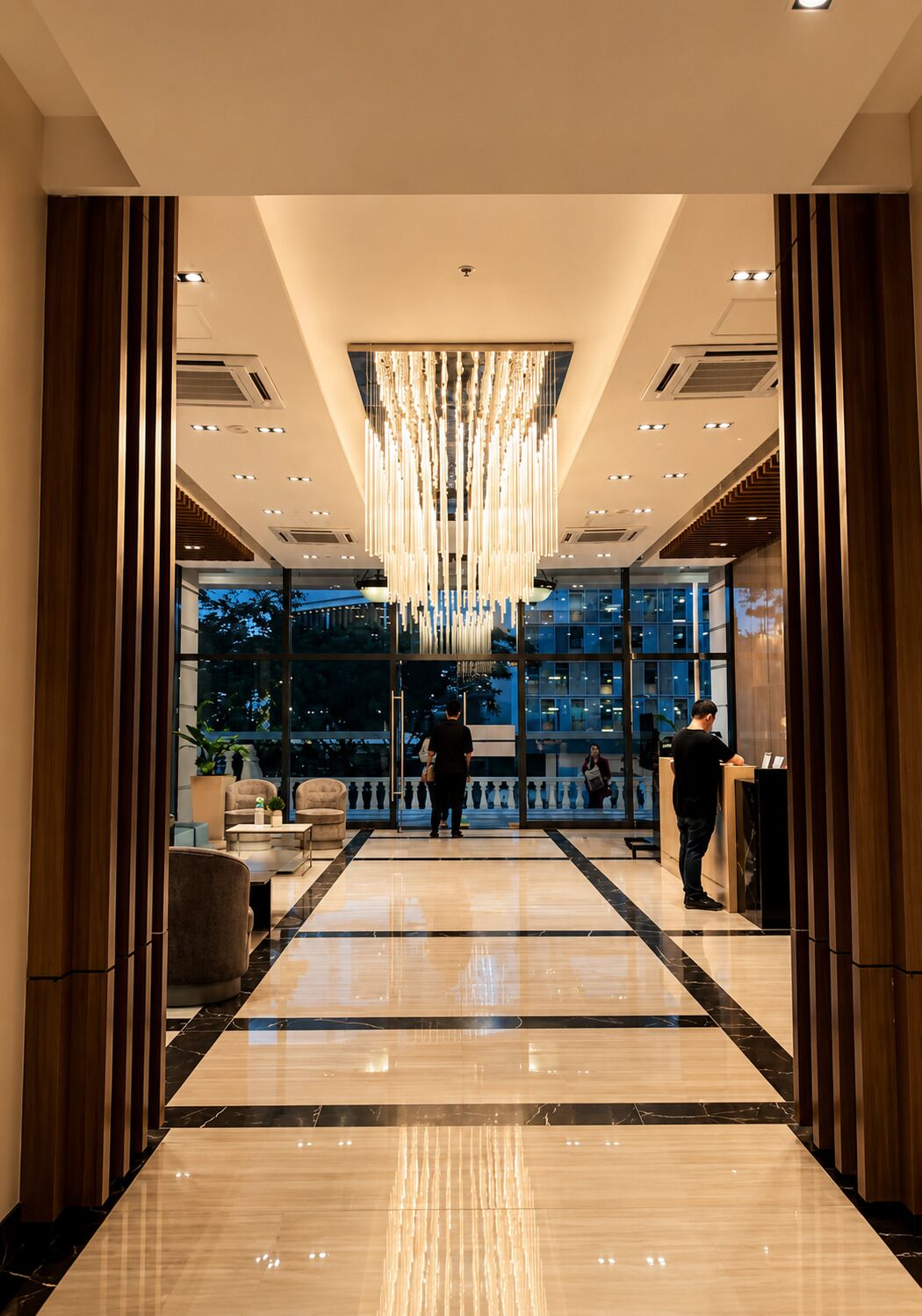 A hotel lobby with a modern chandelier, a reception desk, and a view of the cityscape through large windows.
