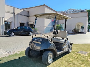 Un véhicule de golf est garé devant un bâtiment, avec une allée pavée et une voiture visible à l’arrière-plan.