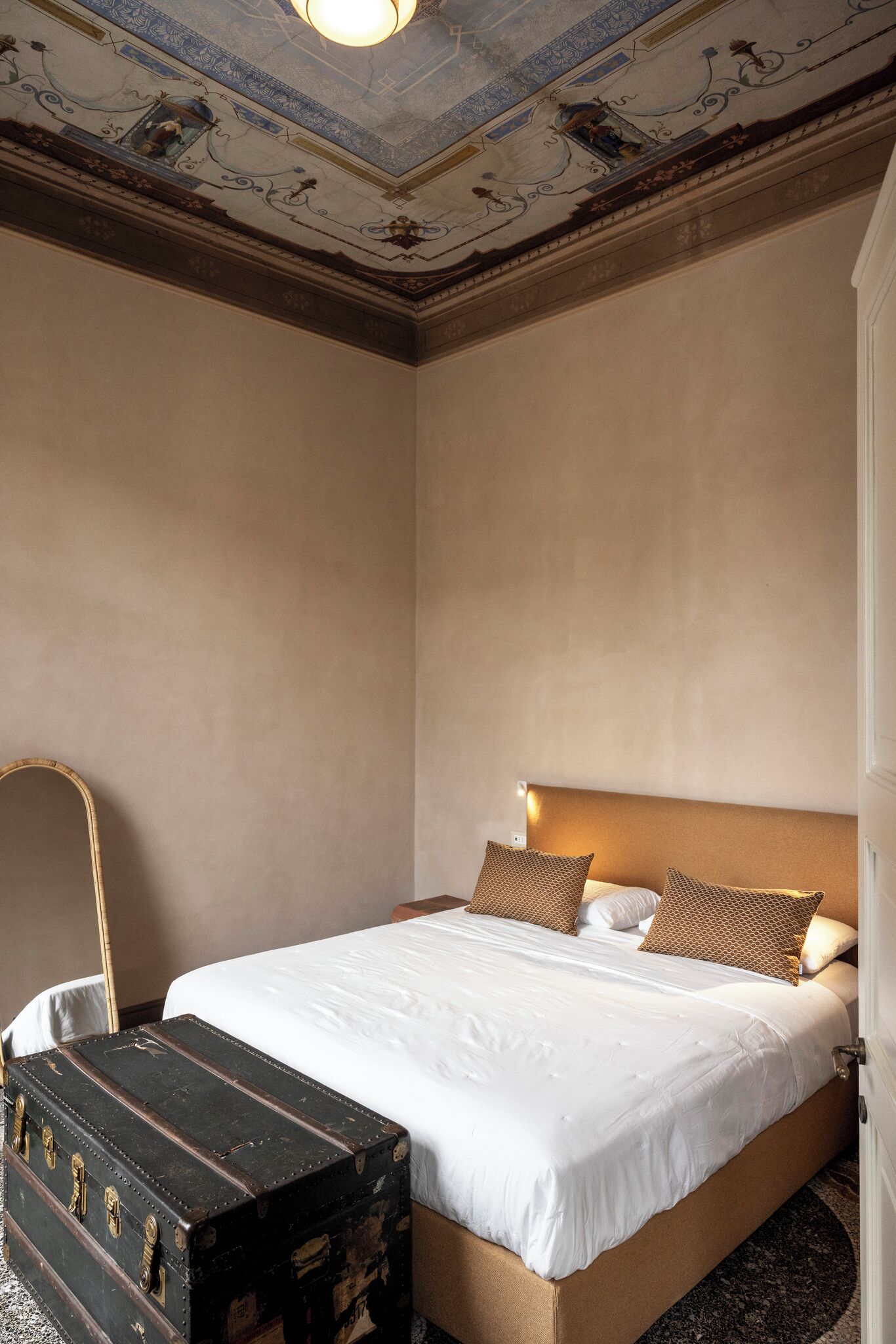 Una camera d'albergo con un letto grande, una testiera in legno, un comodino, una lampada e una valigia d'epoca.