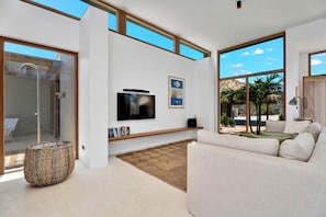 A modern living room with a flat-screen TV, a bookshelf, a large window with a view of palm trees, and a wicker basket. - Brand new luxury Bonaire villa with infinity pool, ocean views, and stunning sunsets. Kas Pajuro accommodates up to 10 guests and offers modern comfort in an exclusive location. (Kralendijk)