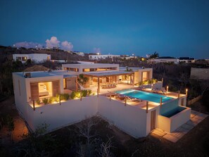A modern multi-story house with a swimming pool, outdoor seating, and a view of the surrounding landscape.