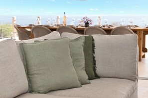 A beige sofa with green and grey cushions in a room with a dining table set for a meal, overlooking a scenic view.