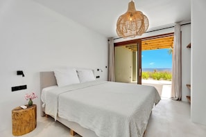 A modern bedroom with a large bed, a wooden side table, a hanging light fixture, and a view of the outdoors. - Brand new luxury Bonaire villa with infinity pool, ocean views, and stunning sunsets. Kas Pajuro accommodates up to 10 guests and offers modern comfort in an exclusive location. (Kralendijk)