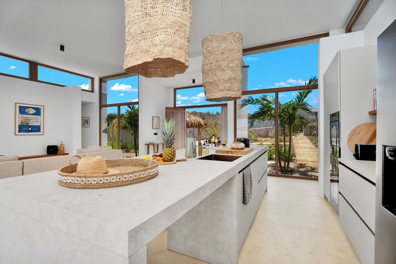 A modern kitchen with a large island, a hanging light fixture, and a view of a tropical landscape.