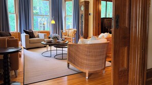 A living room with a sofa, armchair, coffee table, and a wicker chair. There is a clock on the wall and a wooden door. - Heart of Nova Scotia’s South Shore - Curated Luxury in a Landmark Home (Mahone Bay)
