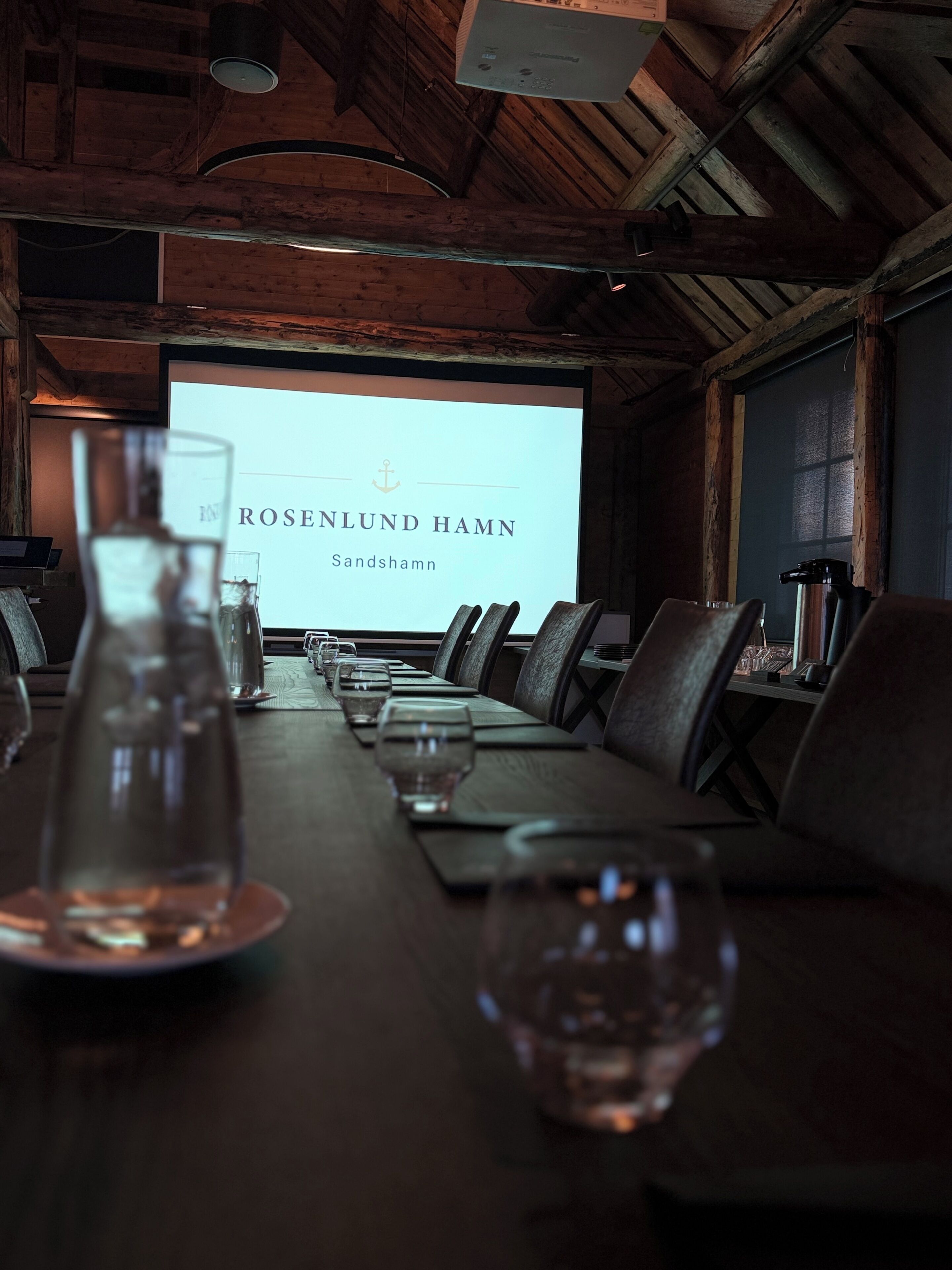 A conference room with a long table set for a meeting, a projector screen displaying 'ROSELUND HAMN Sandshamn', and a wooden ceiling.