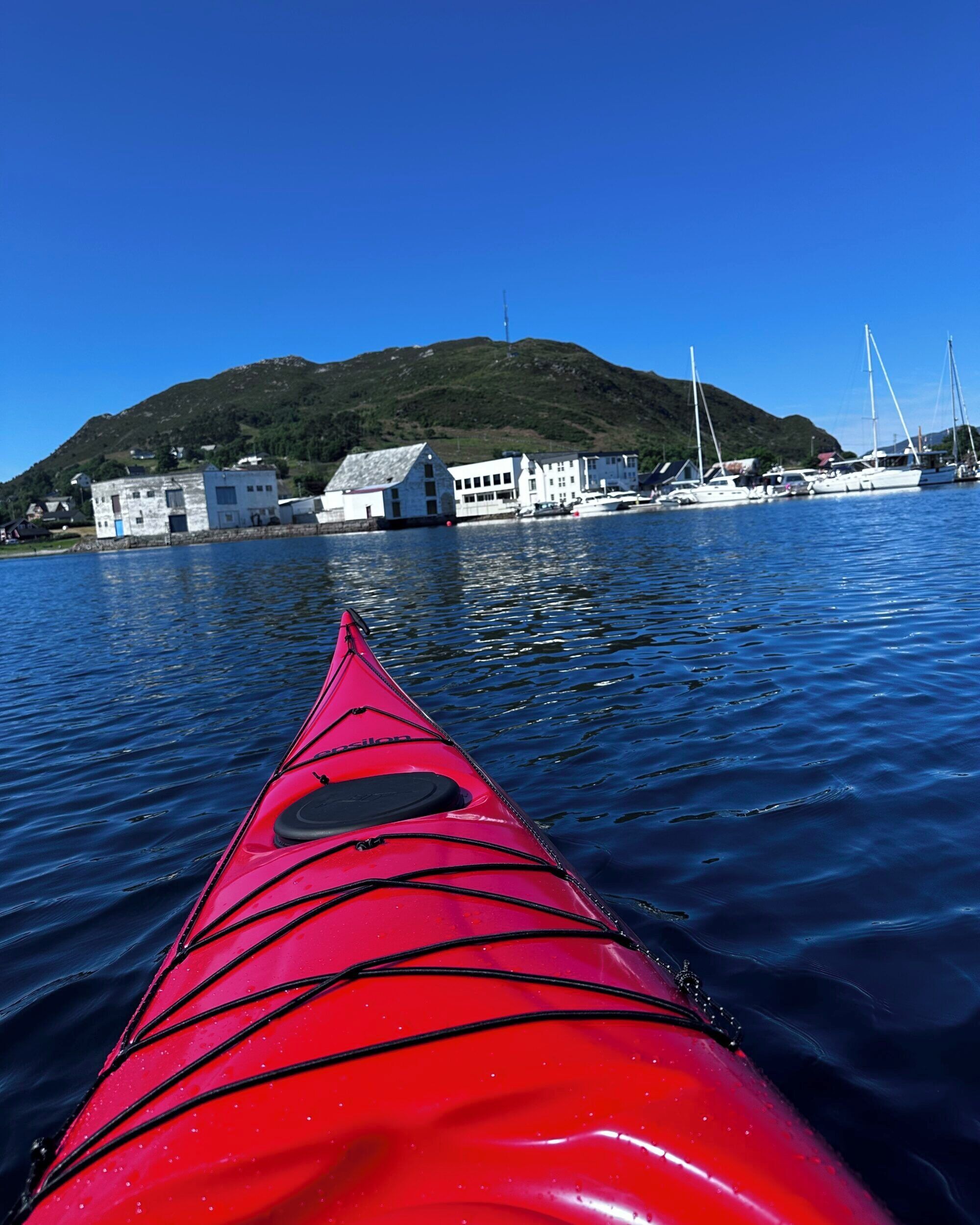 A red kayak on calm water with a view of a hill, buildings, and boats in the background.